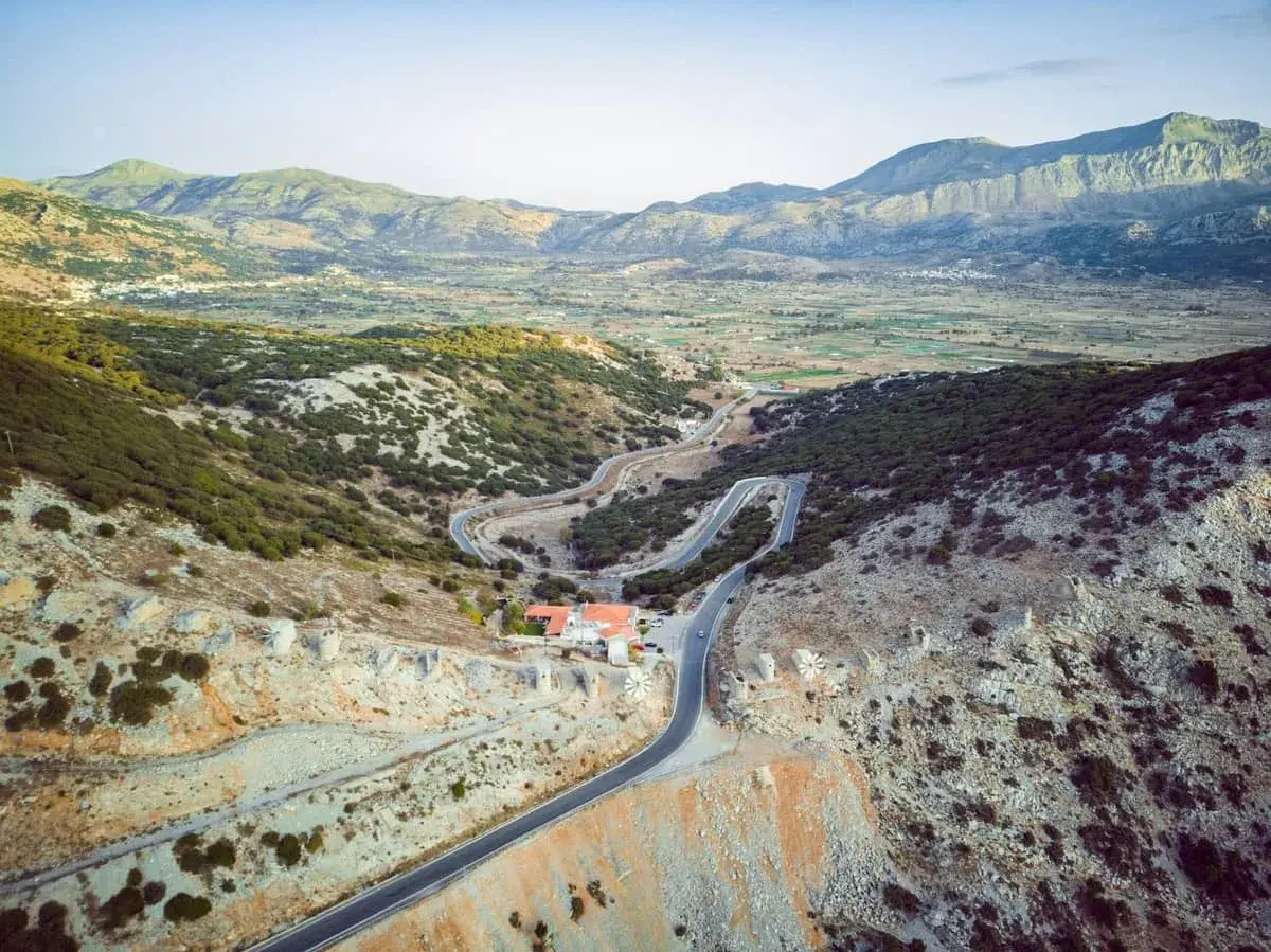 Vue aérienne d'une route sinueuse traversant un paysage montagneux verdoyant, menant à un village sur le plateau de Lassithi. Des ruines de moulins à vent parsèment la colline.