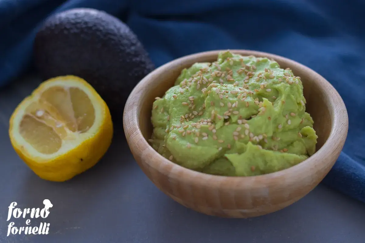 Hummus di avocado cremoso in ciotola di legno, guarnito con semi di sesamo. Accanto, un avocado intero e mezzo limone.