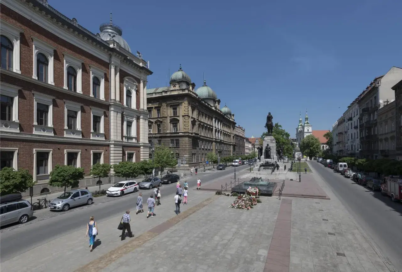 Kraków: pomnik Grunwaldzki, zabytkowe budynki i spacerujący ludzie. Wśród nich można sobie wyobrazić znane osoby z Krakowa.