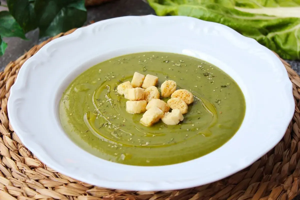 Crema de acelgas verde vibrante, servida en plato blanco con picatostes dorados y un chorrito de aceite de oliva.
