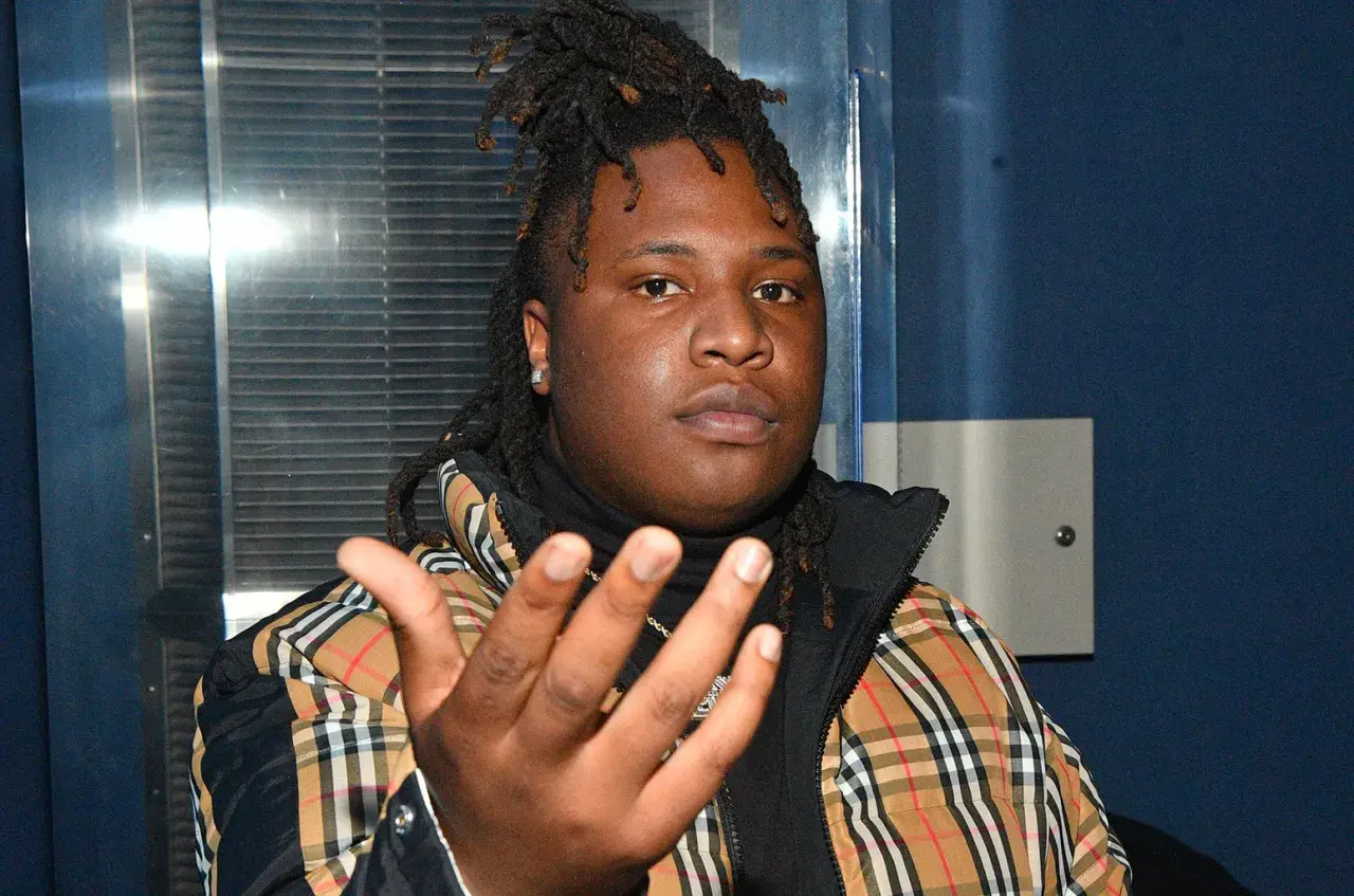 A young man with dreadlocks, wearing a plaid jacket, gestures forward. He's the artist behind many popular Tay Keith songs.