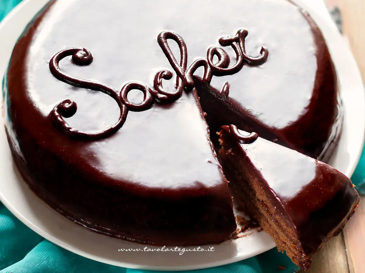 Una lucida torta viennese al cioccolato, con una fetta tagliata, decorata con la scritta "Sacher" in glassa scura.