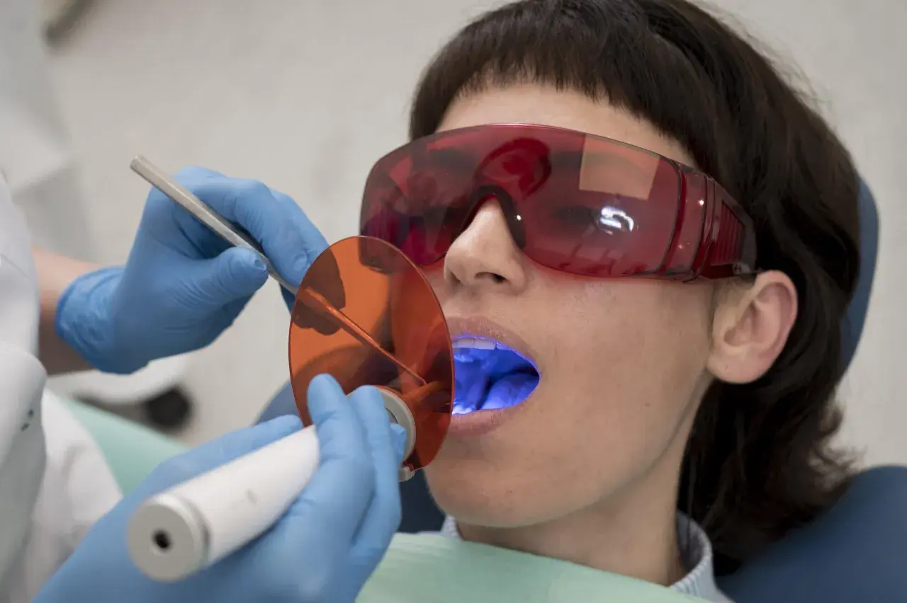 Pacjentka w okularach ochronnych poddaje się zabiegowi stomatologicznemu. Dentysta używa lampy polimeryzacyjnej, która emituje niebieskie światło. Po piaskowaniu zębów, taka kawa z mlekiem smakuje jak nagroda.