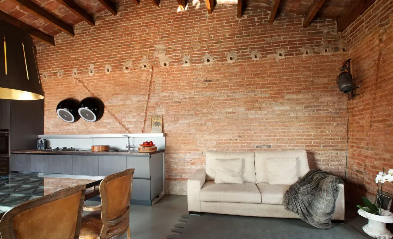 Cocina y sala de estar con paredes de ladrillo visto, un sofá beige y sillas de cuero. El acabado rústico en paredes aporta calidez.