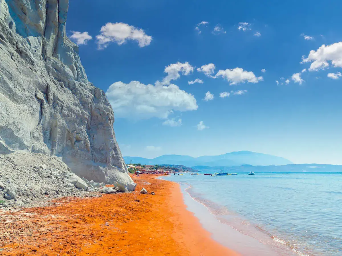 Ein atemberaubender Kefalonia Strand mit leuchtend orangefarbenem Sand, der von einer steilen weißen Klippe eingerahmt wird. Das türkisfarbene Meer lädt zum Schwimmen ein.