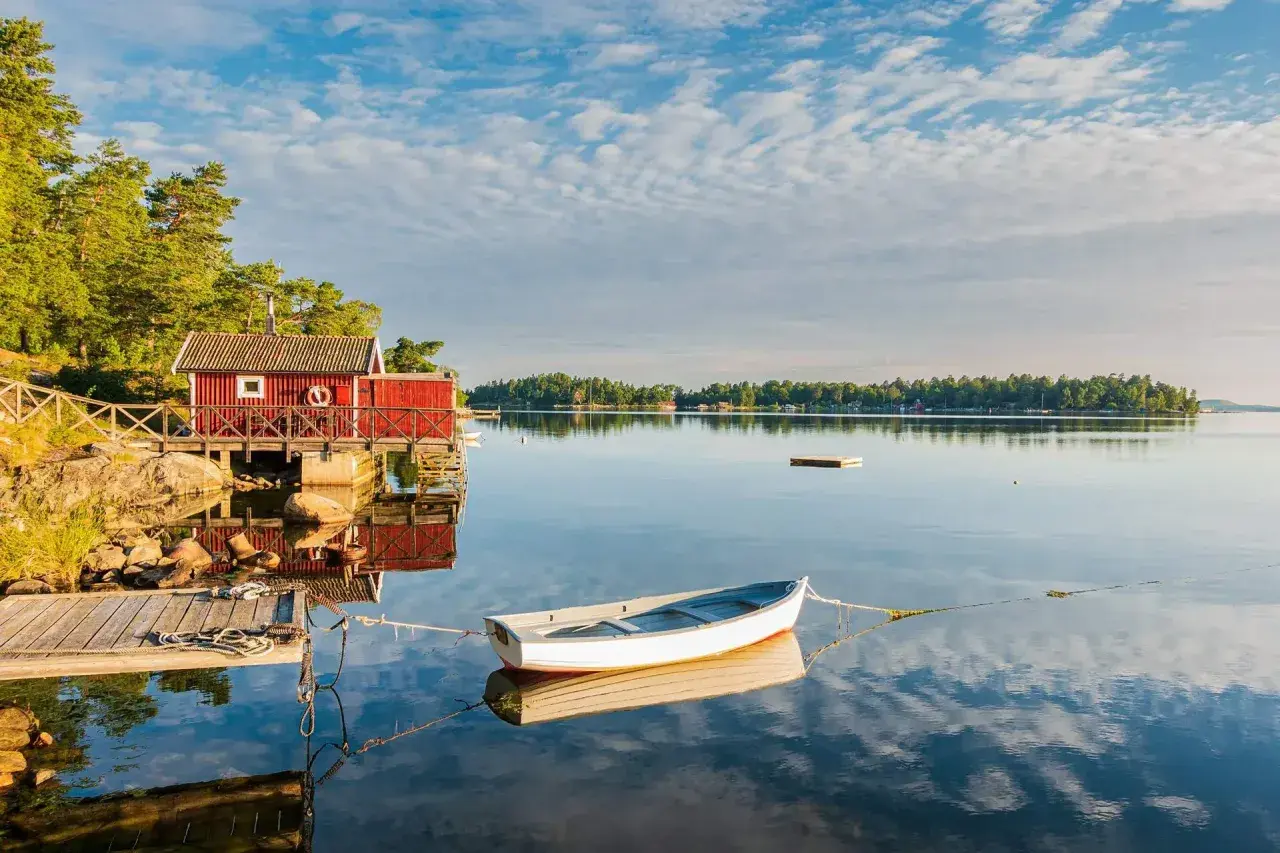 En vit båt ligger förtöjd vid en röd sjöbod. Kanske är det en båt till Drottningholm, redo för en lugn tur på vattnet.