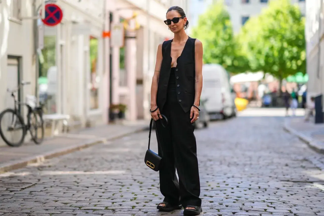 Eine Frau trägt ein festliches Oberteil zu schwarzer Hose, Sonnenbrille und einer kleinen schwarzen Tasche.
