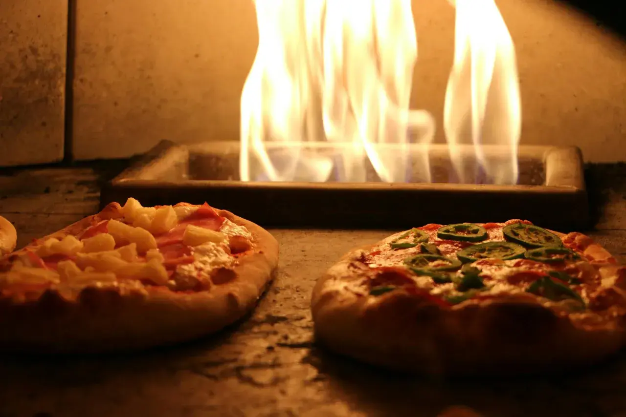 Deux pizzas maison, une à l'ananas et jambon, l'autre aux poivrons verts, cuisent dans un four avec des flammes vives. Une recette pizza maison avec pâte toute prête facile et rapide.