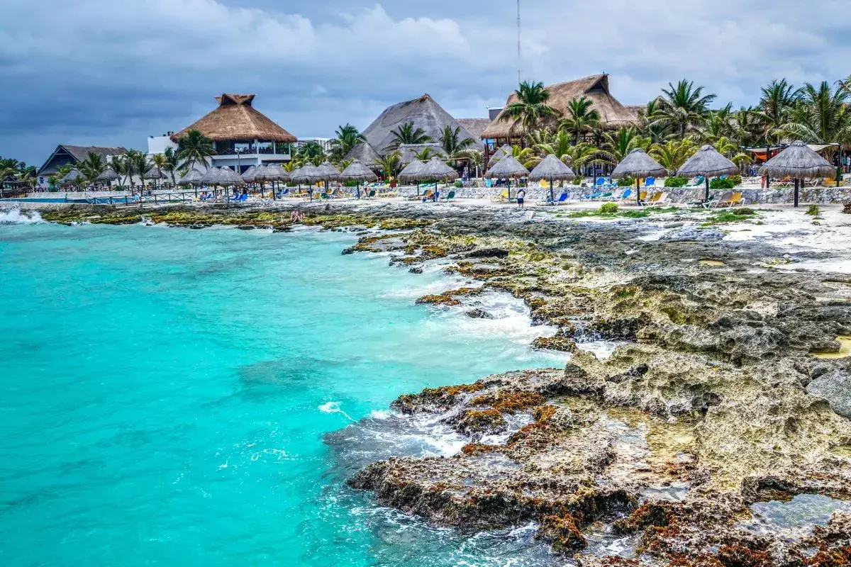 Plage mexicaine avec eau turquoise, rochers, palmiers et bungalows à toit de chaume.