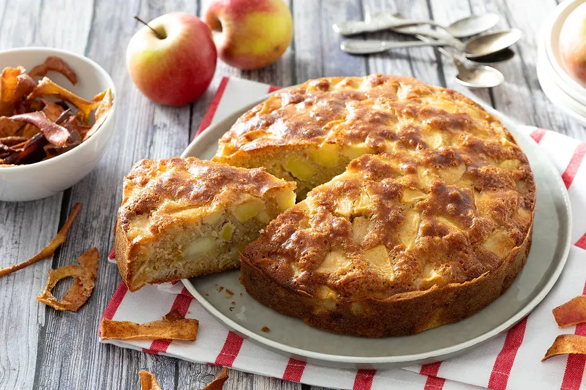 Un délicieux gâteau aux pommes, doré et moelleux, avec des morceaux de fruits visibles. Des pommes fraîches et des chips de pommes complètent cette scène gourmande.