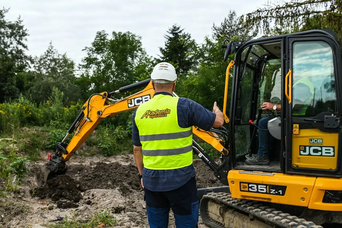 Instruktor pokazuje, jak szybko można ukończyć kurs na koparkę. Operator w kamizelce "Akademia Operatora" uczy się obsługi maszyny JCB.