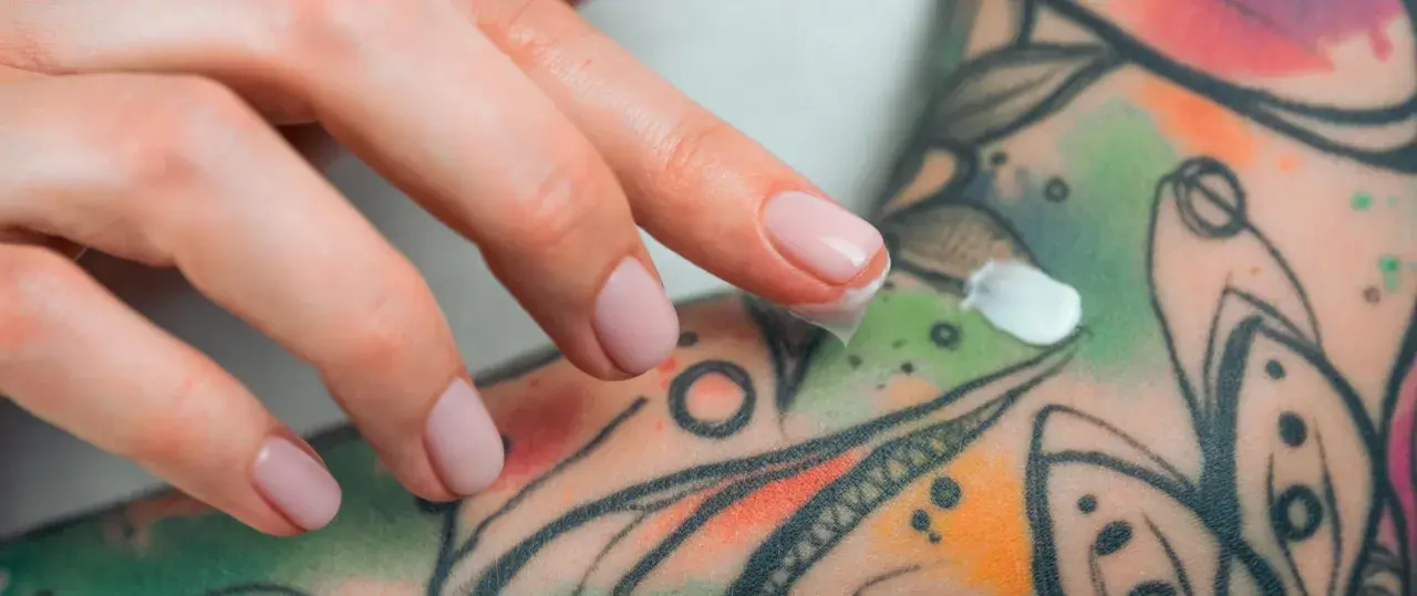 Main d'une personne appliquant une crème sur un tatouage coloré. Si vous suspectez une infection tatouage, que faire ? Consulter un professionnel de santé.
