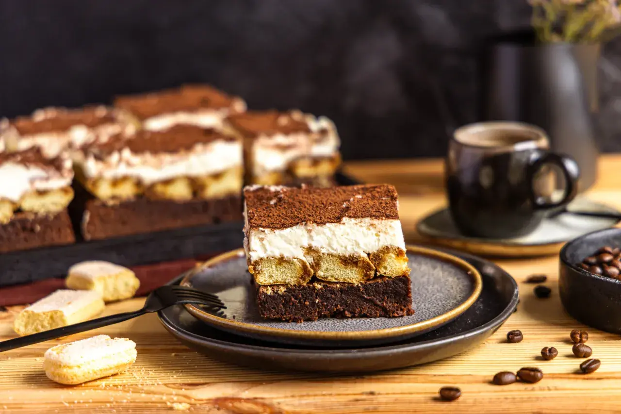 Ein Stück Tiramisu Brownie mit Löffelbiskuits, cremiger Füllung und Kakaopulver. Daneben eine Tasse Kaffee und Kaffeebohnen.
