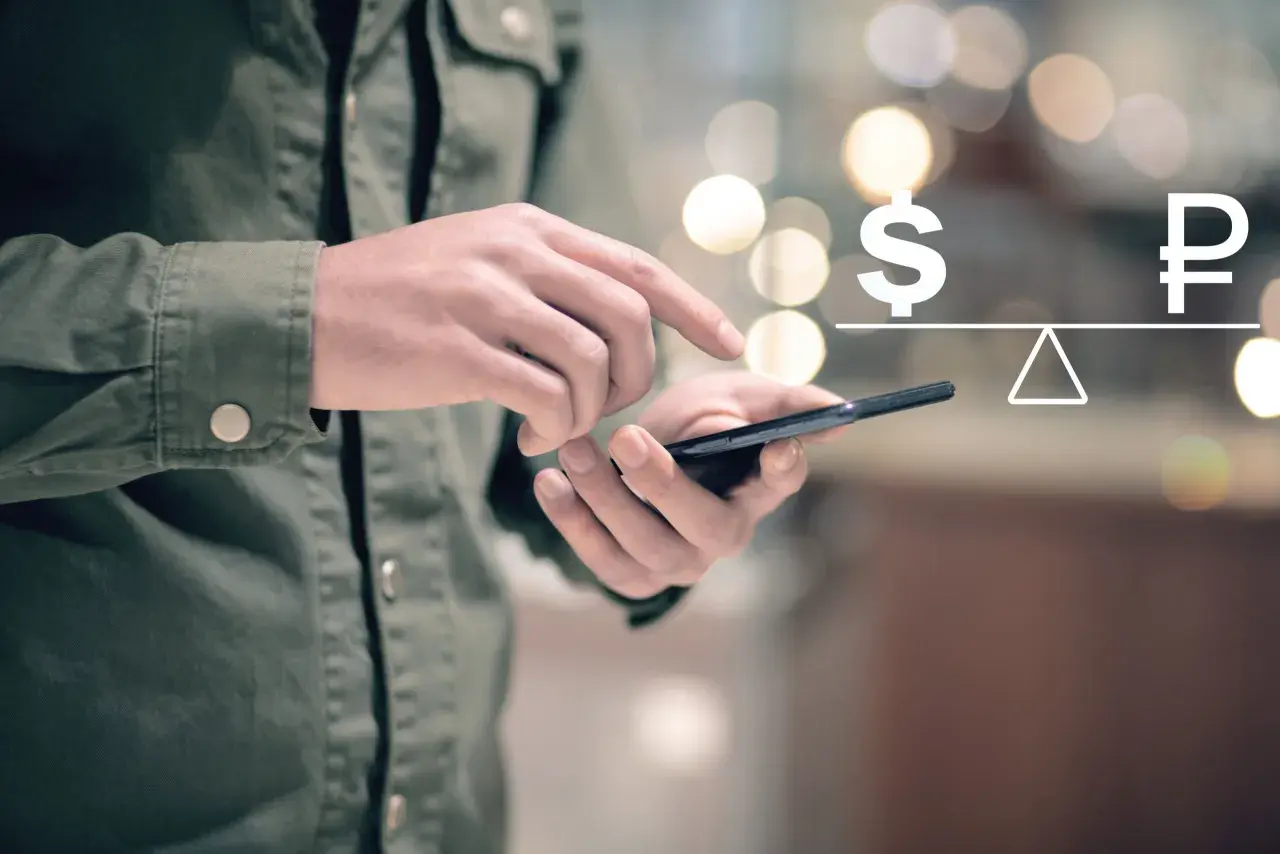 Dłoń trzyma telefon, na ekranie symbol dolara i rubla na wadze. Symbolizuje to paypal przewalutowanie.