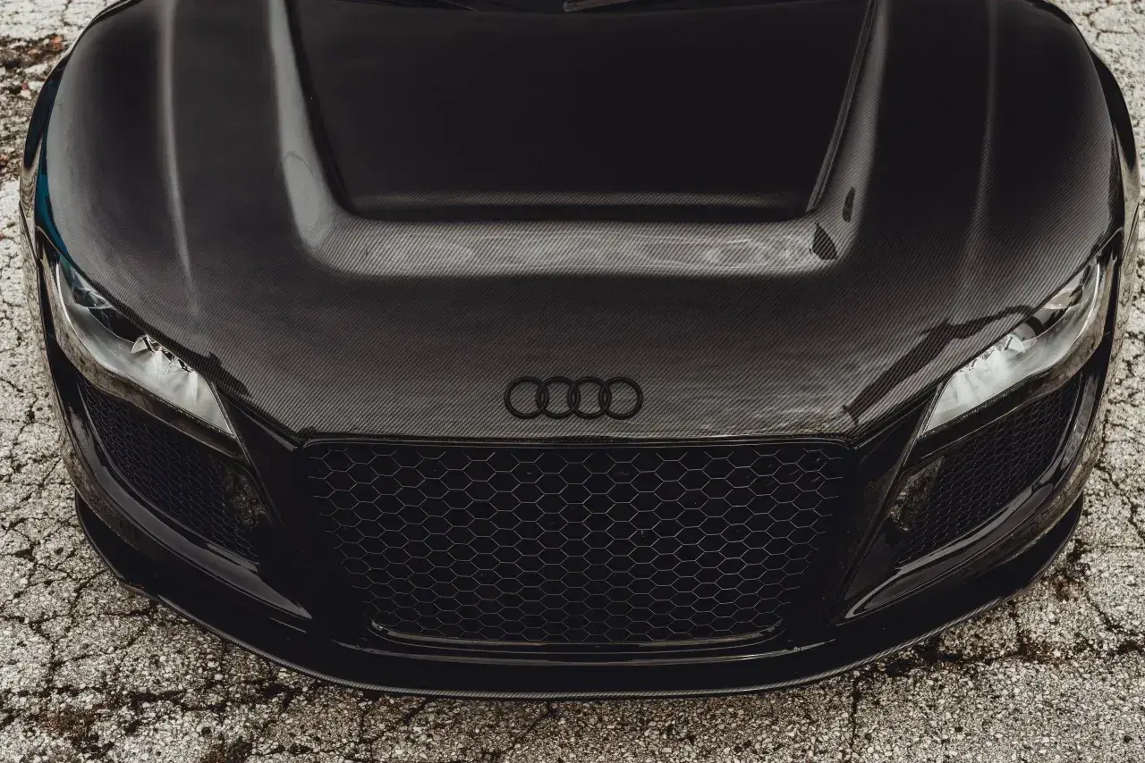 Przód czarnego auta sportowego z maską z włókna węglowego. Widać reflektory, grill i logo Audi.