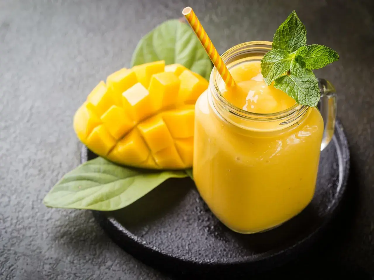 Un smoothie mangue onctueux dans un pot en verre, garni de menthe fraîche, à côté d'une moitié de mangue coupée en dés.