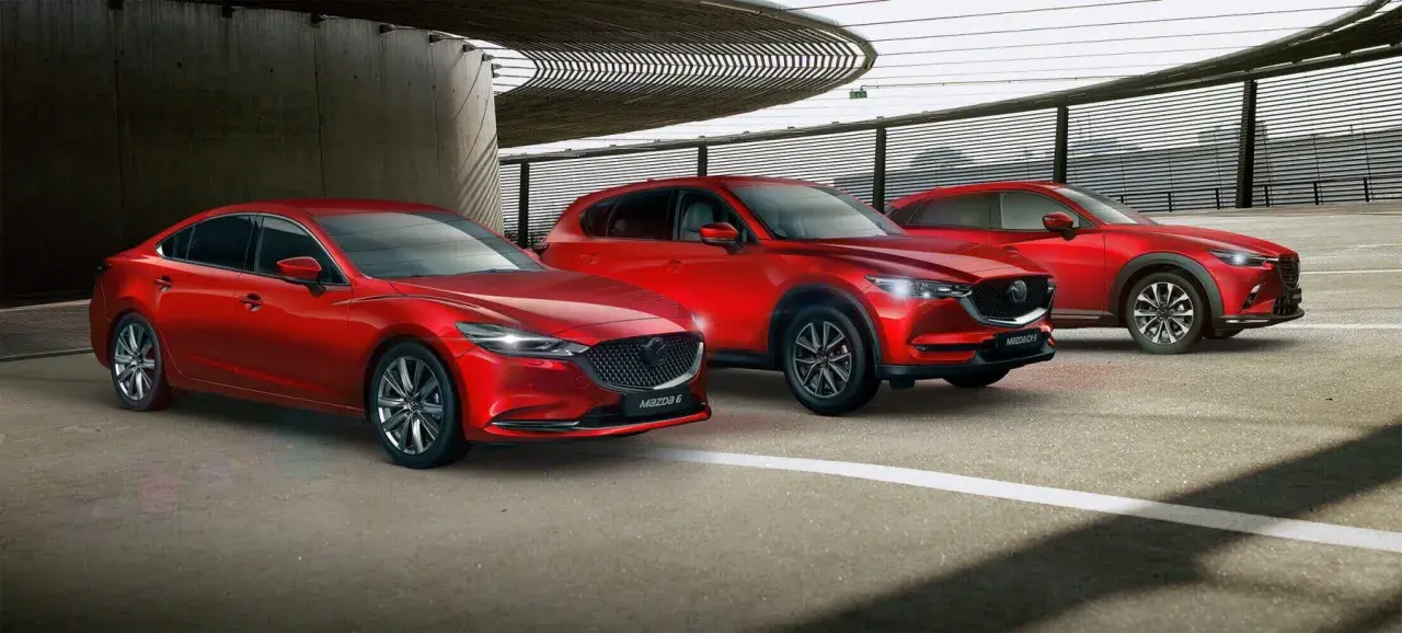 Trzy czerwone samochody marki Mazda: sedan, SUV i crossover, zaparkowane pod nowoczesną konstrukcją.