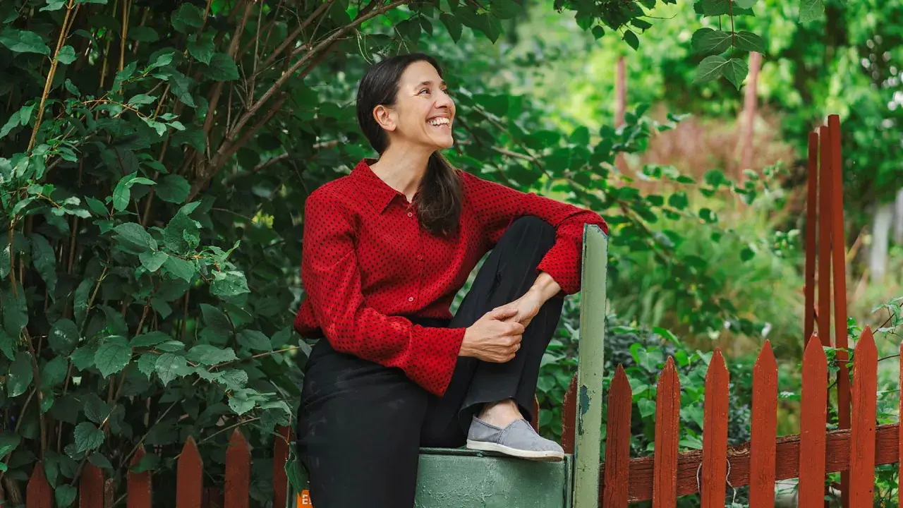 Paloma Grandin sitter avslappnat vid ett staket, omgiven av grönska. Hon ler och tittar uppåt, fångad i ett ögonblick av glädje.