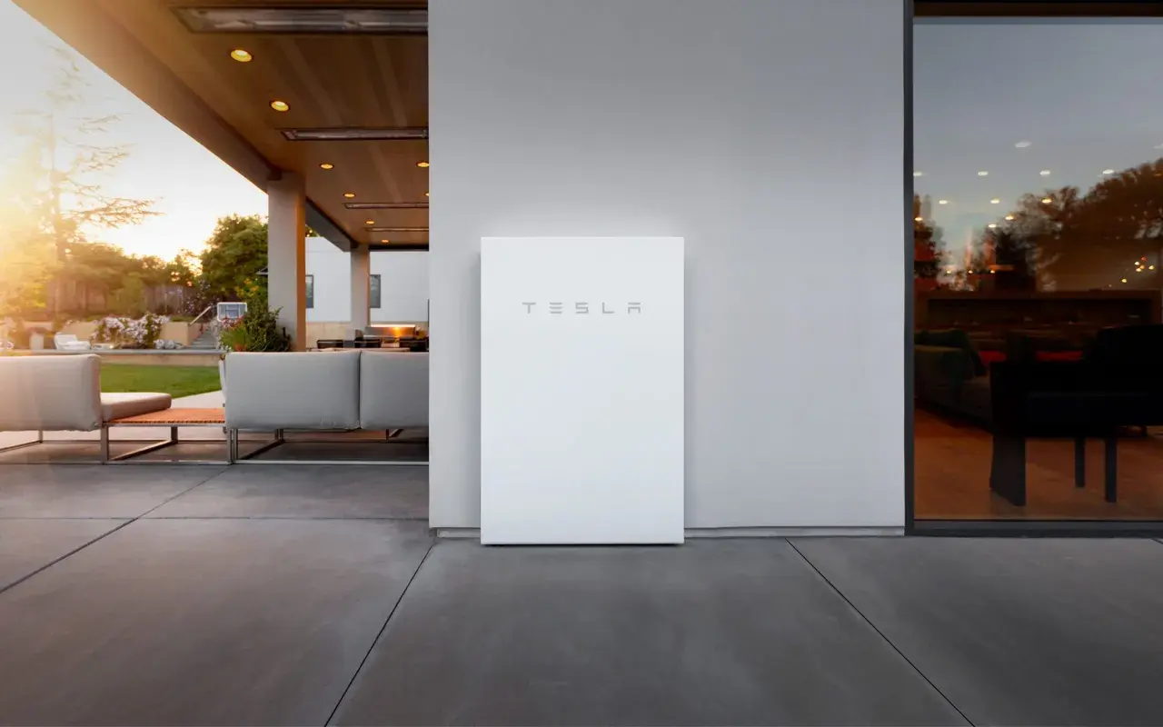 Un elegante Tesla Powerwall blanco se alza junto a una casa moderna con un patio y muebles de exterior.