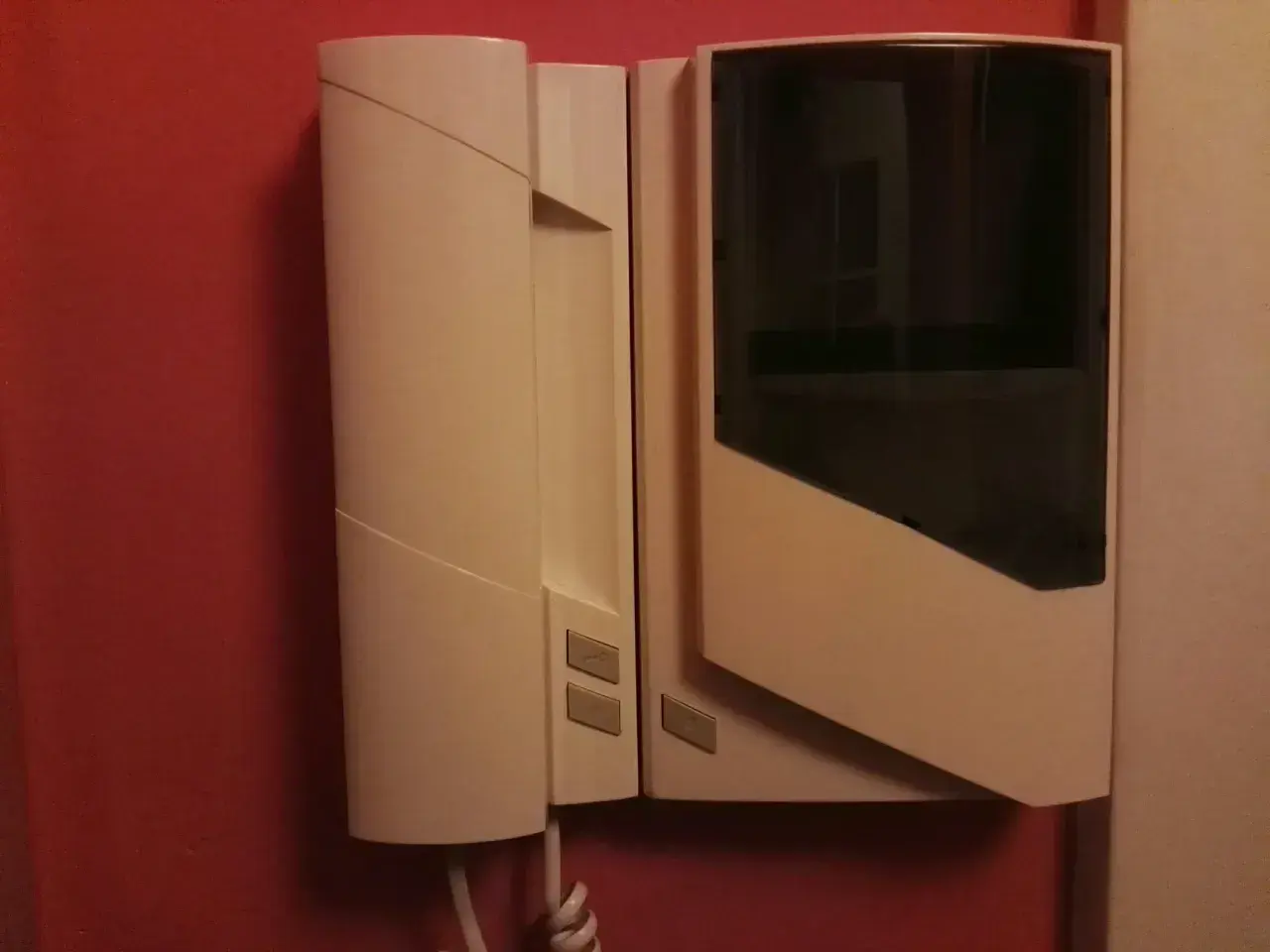 Biały unifon zamontowany na czerwonej ścianie. Urządzenie ma prosty, nowoczesny design z ciemną szybką.