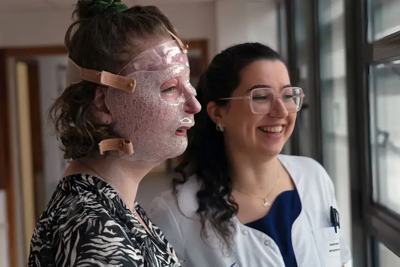 Une femme porte un masque transparent sur le visage, peut-être pour aider à comment cacher une brûlure. Une autre femme, souriante, la regarde.