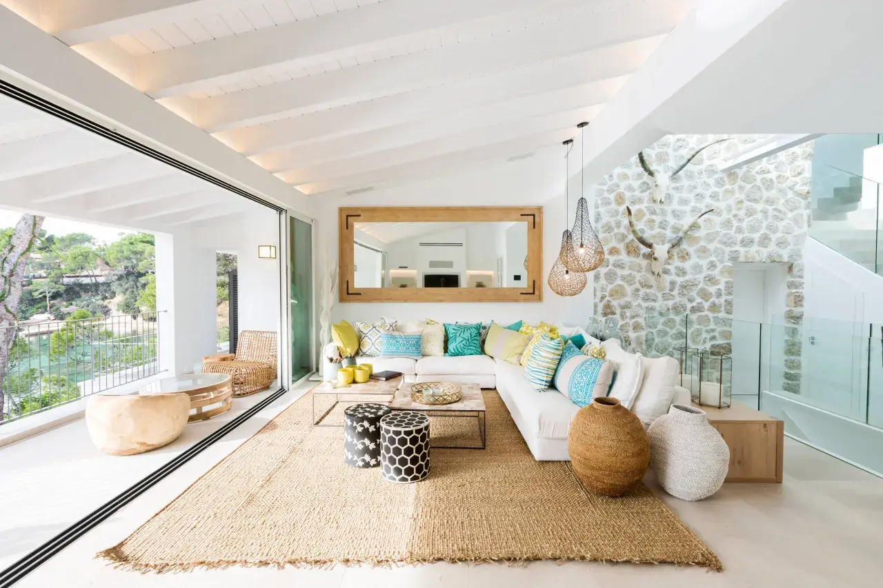 Helles Wohnzimmer mit Finca Style Einrichtung, weißem Sofa, bunten Kissen, Jute-Teppich und Blick auf das Meer.
