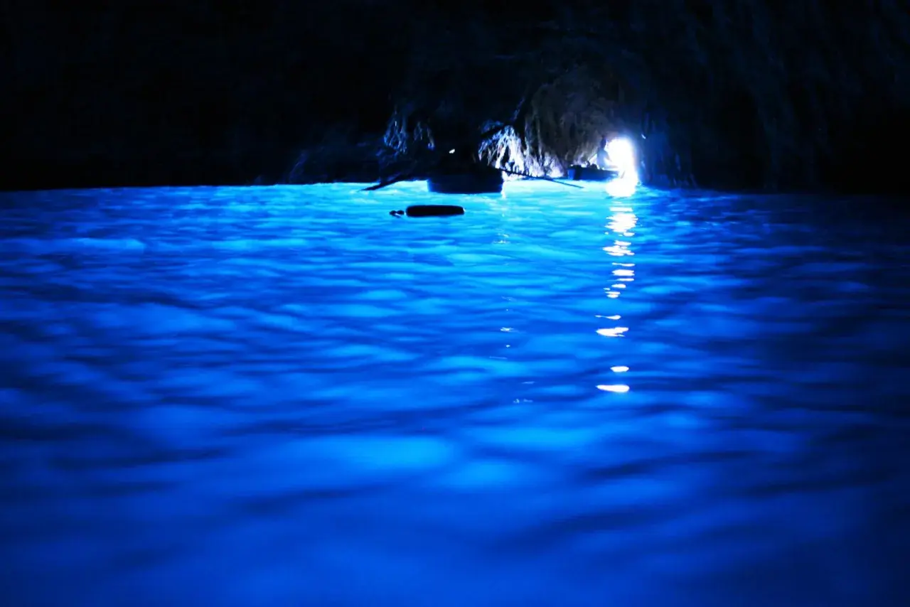 Acqua blu elettrico nella grotta, un piccolo remo galleggia mentre una barca si dirige verso la luce all'uscita della blaue grotte.