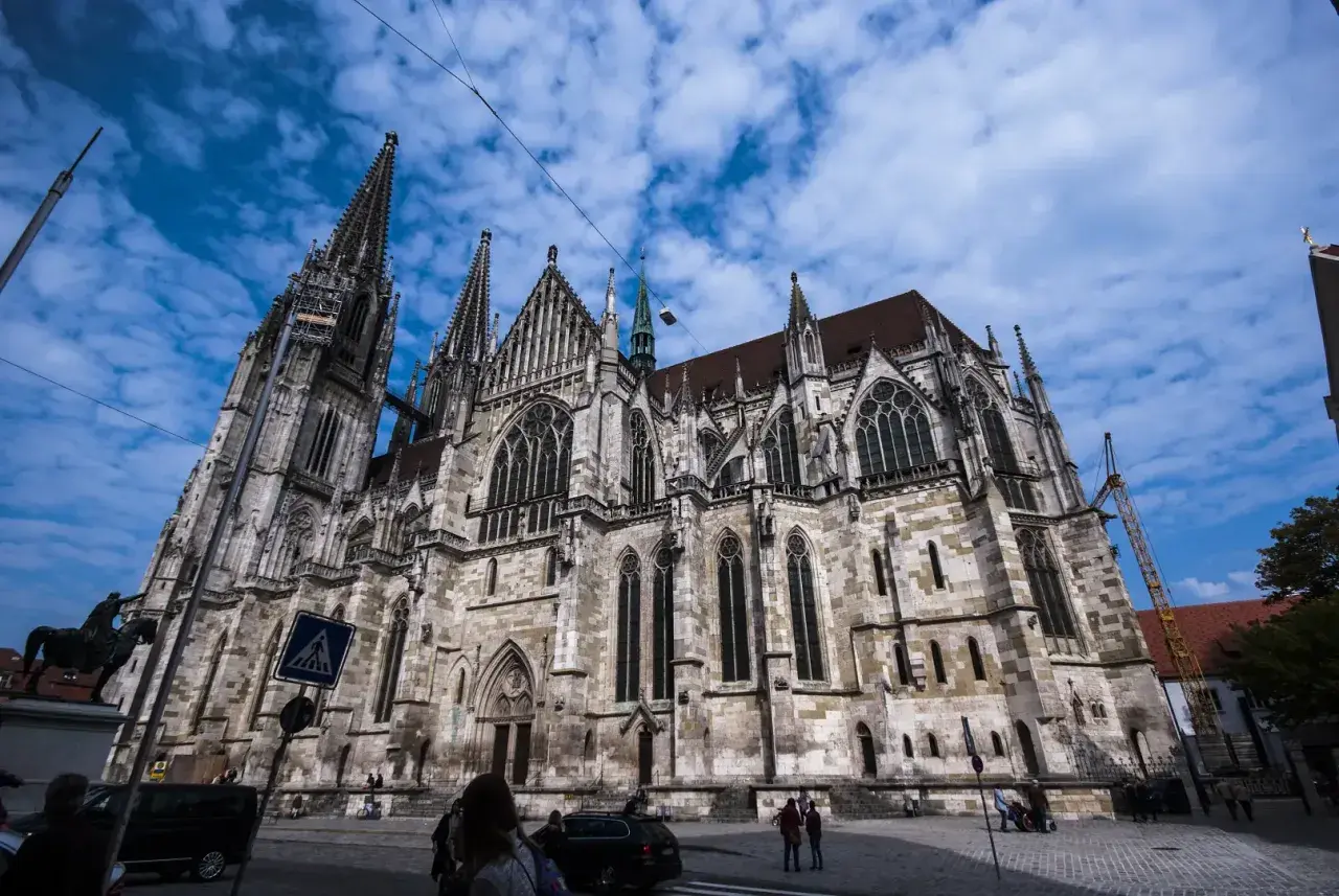 Der imposante Dom Regensburg ragt mit seinen Türmen in den blauen Himmel. Menschen und Autos bewegen sich auf dem Platz davor.