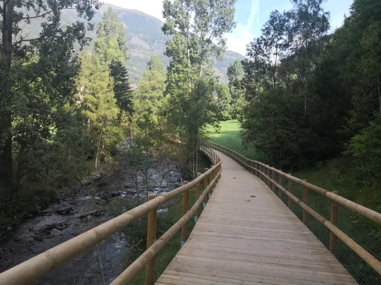 Paseo de madera junto a un río, parte de la ruta del ferro andorra, rodeado de árboles y montañas.