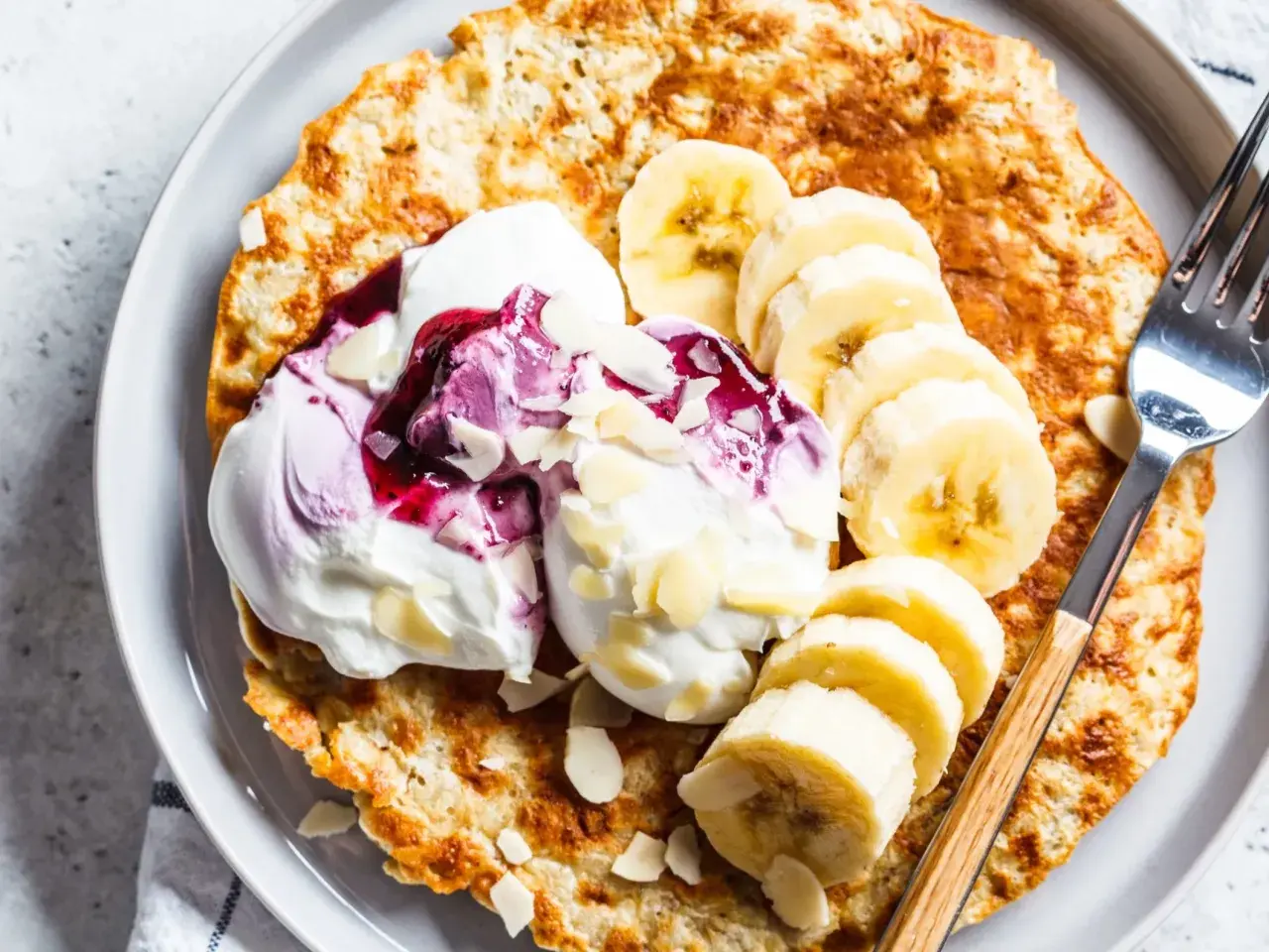 Puszysty omlet bananowy z jogurtem, musem jagodowym i płatkami migdałów na talerzu.