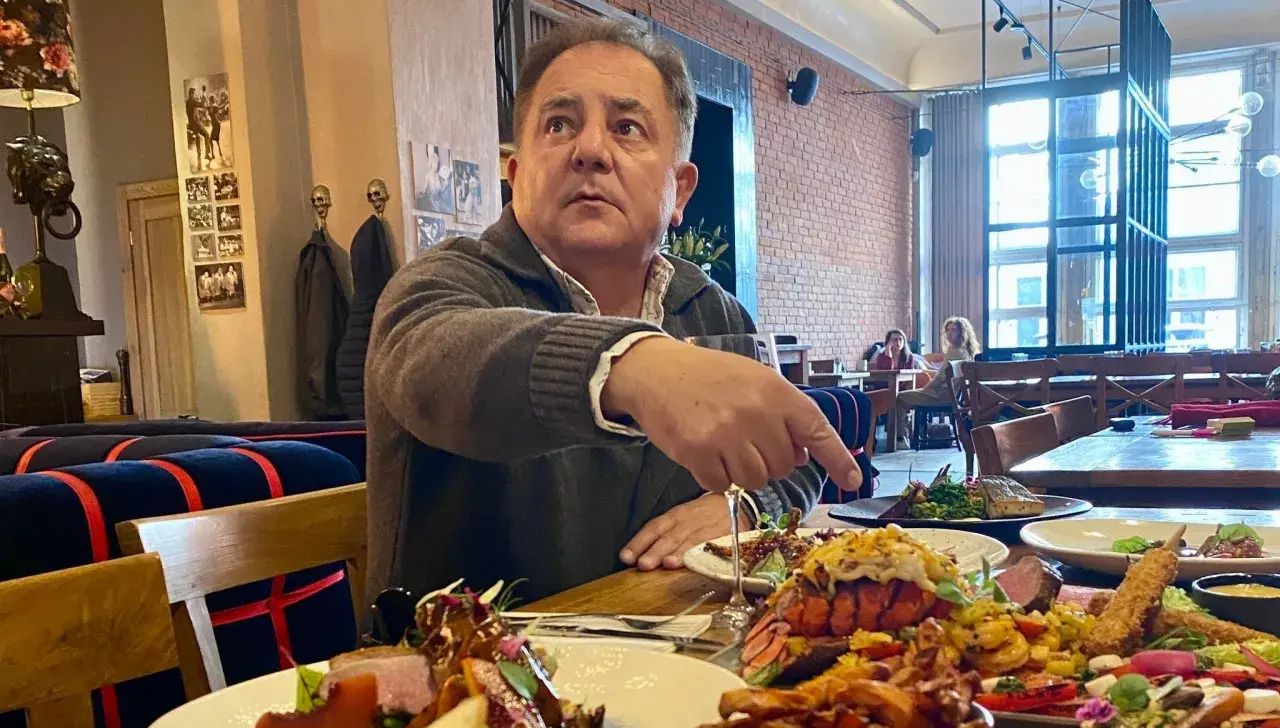 Robert Makłowicz wskazuje na obficie zastawiony stół w restauracji. Widać dania z owocami morza i mięsem.
