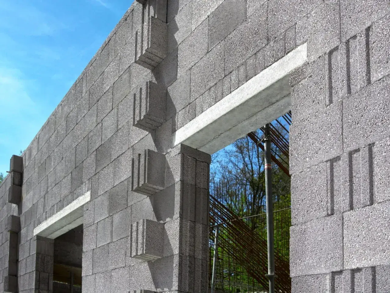 Ściana z bloczków betonowych z widocznym betonowym nadprożem nad otworem okiennym. W tle zbrojenie i drzewa.