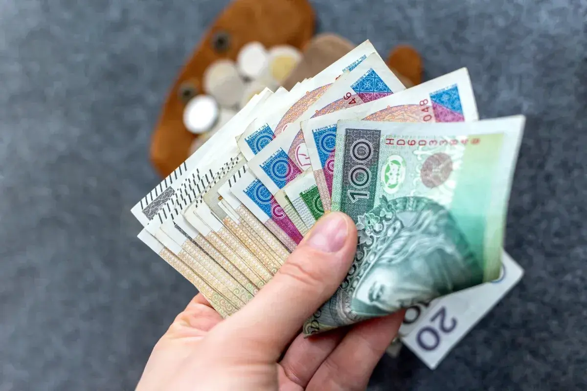 Dłoń trzymająca wachlarz polskich banknotów, w tle portfel z monetami.