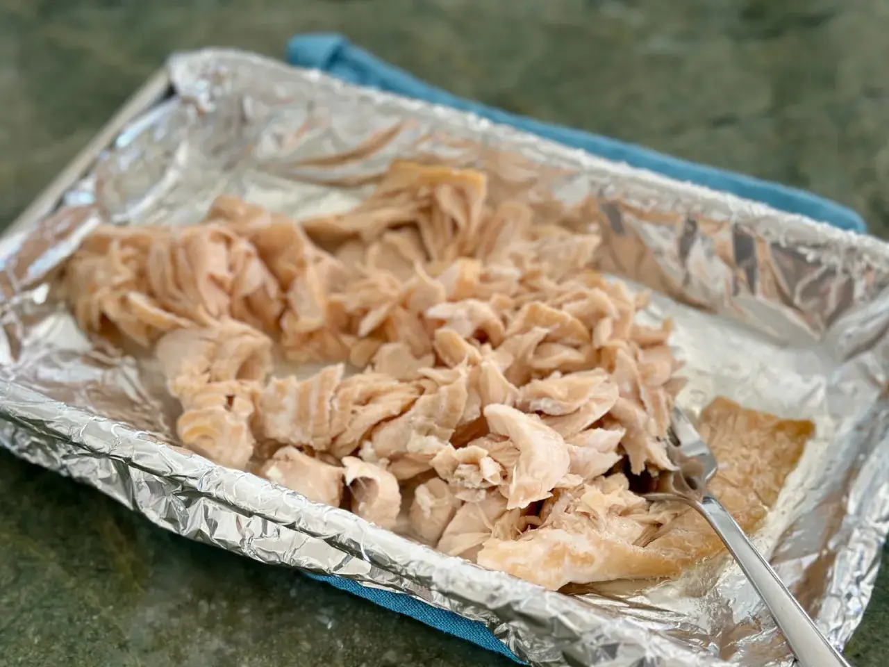 Flaked cooked salmon in a foil-lined baking dish, ready for a salmon flakes recipe. A fork rests on the side.