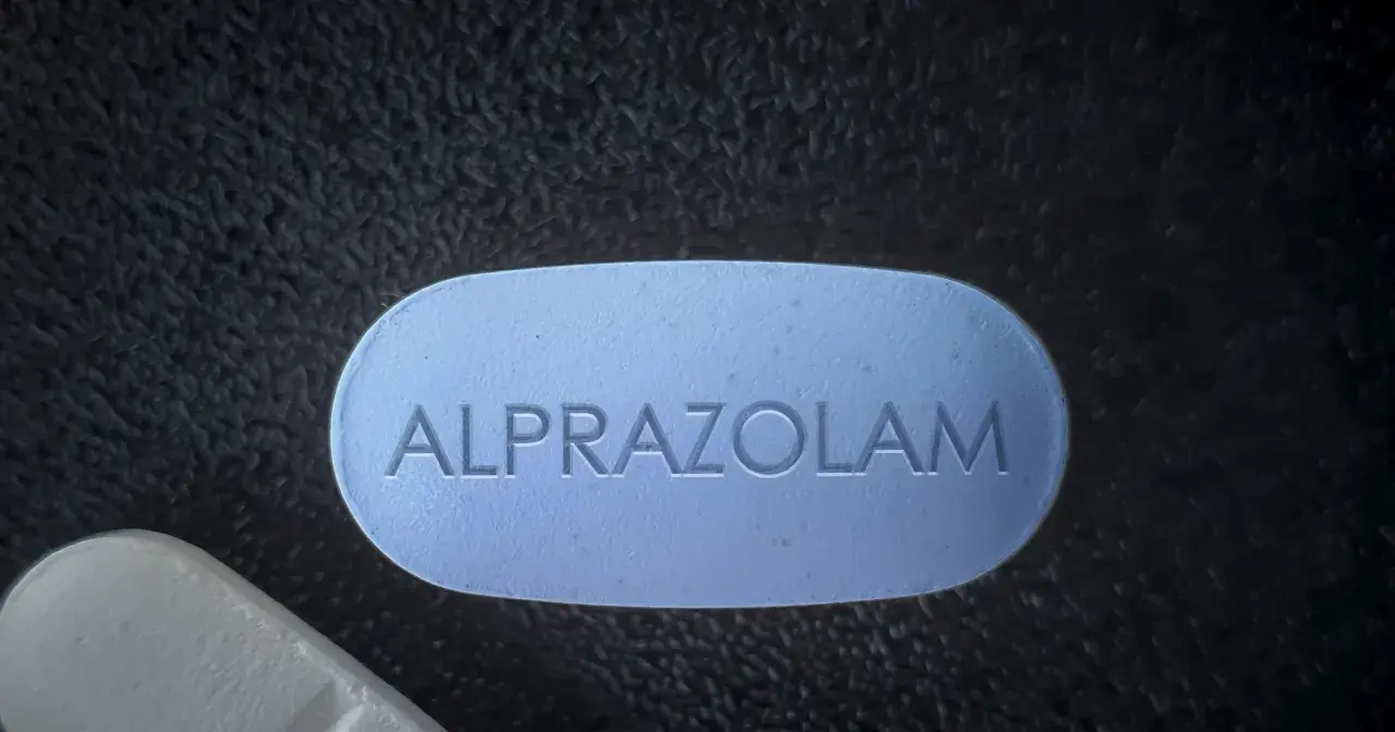 Niebieska tabletka z napisem ALPRAZOLAM leży obok białej tabletki na ciemnym, teksturowanym tle.
