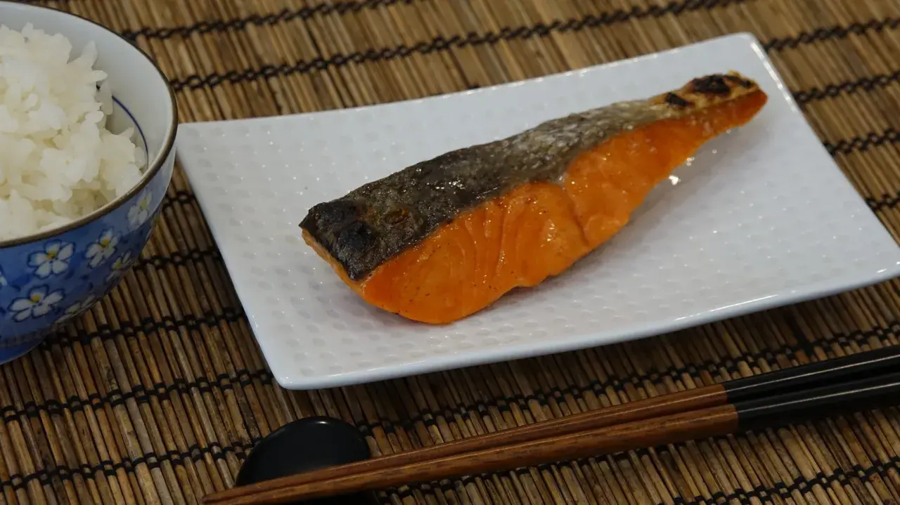 A perfectly grilled salmon shioyaki rests on a white plate, next to a bowl of white rice and chopsticks.