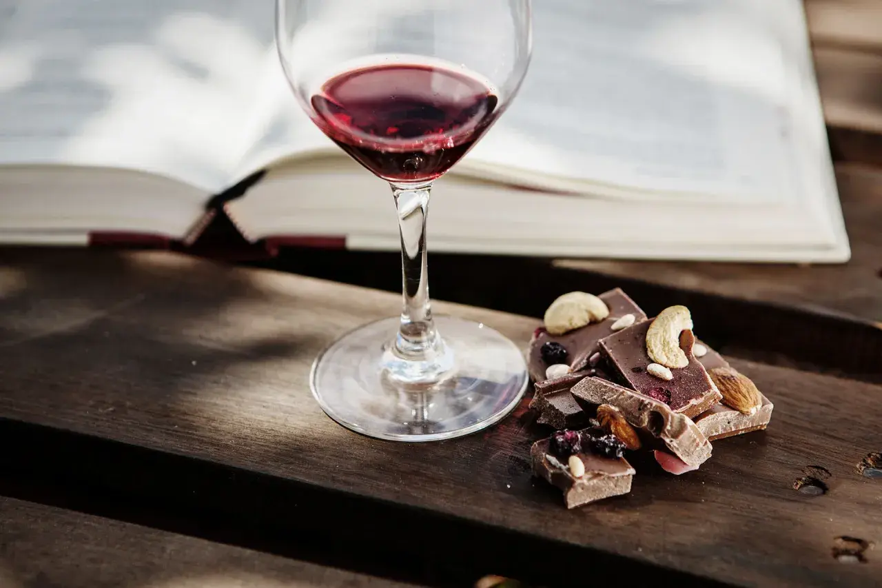 Un bicchiere di vino rosso accanto a pezzi di cioccolato con frutta secca e noci. Un perfetto abbinamento vino e cioccolato per una serata di relax.