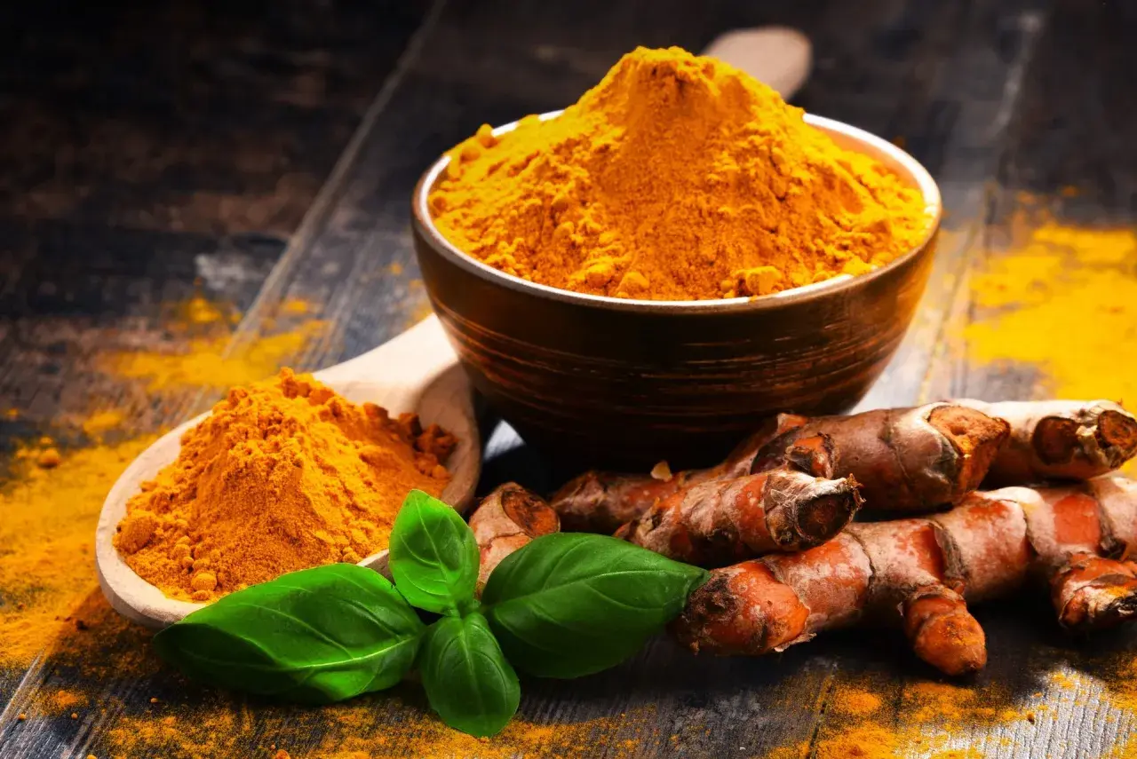 Radici di curcuma e polvere dorata in un cucchiaio e ciotola, con foglie verdi. La curcuma è nota per le sue proprietà antinfiammatorio.