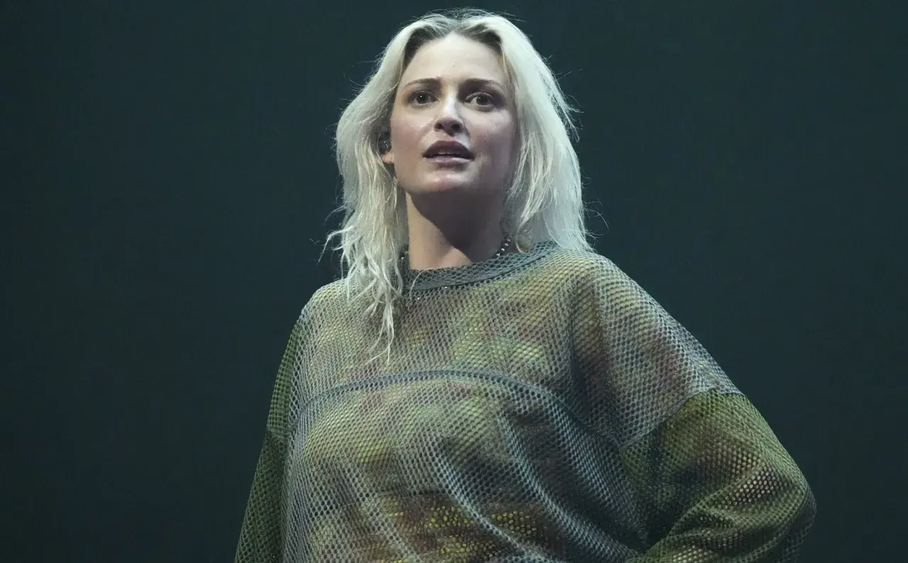 Blond wokalistka w siatkowej bluzce, przypominająca styl wokalisty Linkin Park, na ciemnym tle.