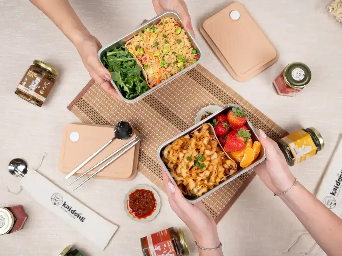 Two hands hold separate noodle bento boxes, one with fried rice and greens, the other with noodles, strawberries, and orange slices.