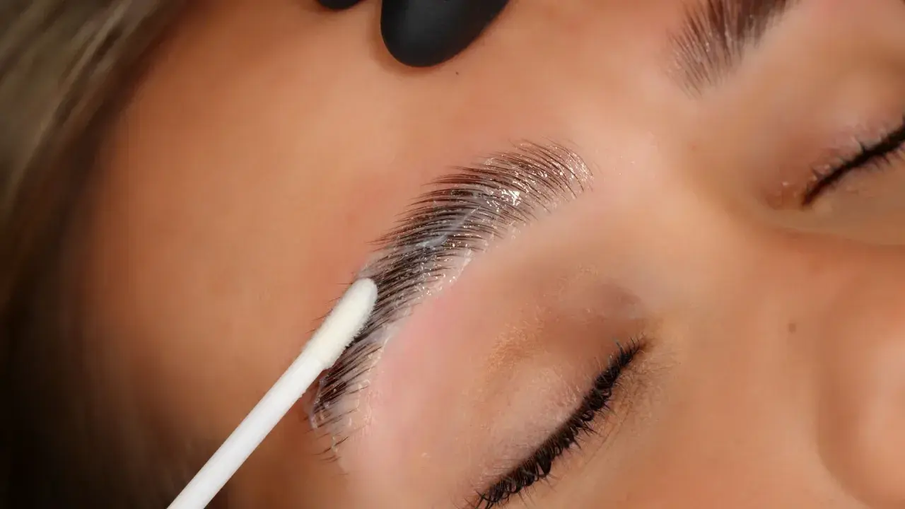 Aplicación de producto para que es el laminado de cejas. Un hisopo blanco aplica una crema sobre las cejas oscuras y peinadas.