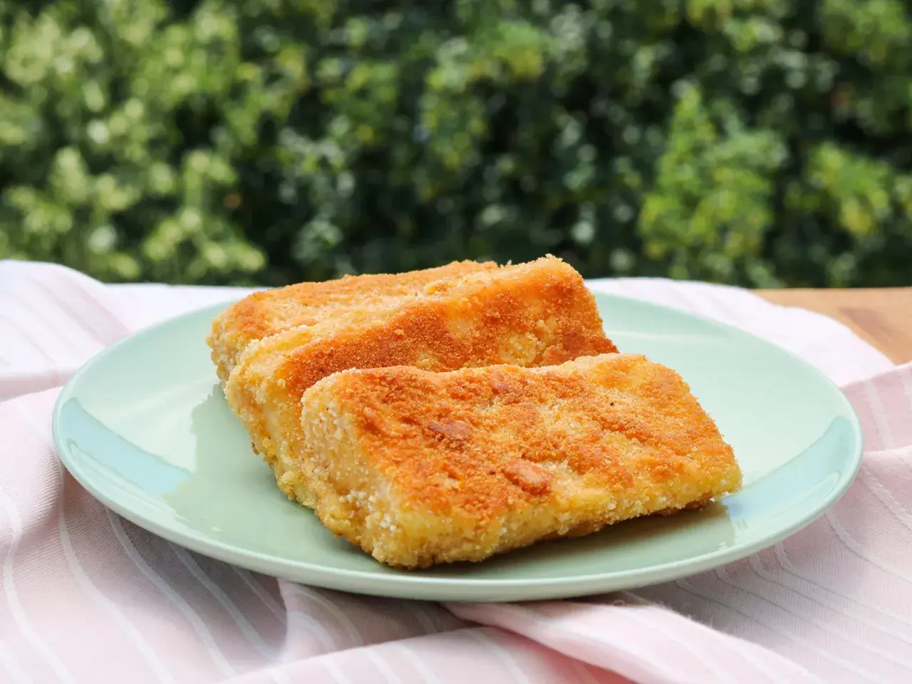 Zwei goldbraun gebackene Emmentaler-Schnitzel auf einem hellgrünen Teller, serviert mit einem rosafarbenen Tuch.