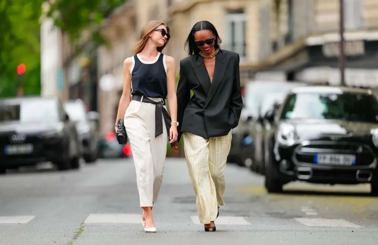 Deux femmes marchent dans la rue. L'une porte un pantalon blanc et un top noir, l'autre un blazer noir et un pantalon rayé. Idées pour comment s'habiller quand on est mince et petite.