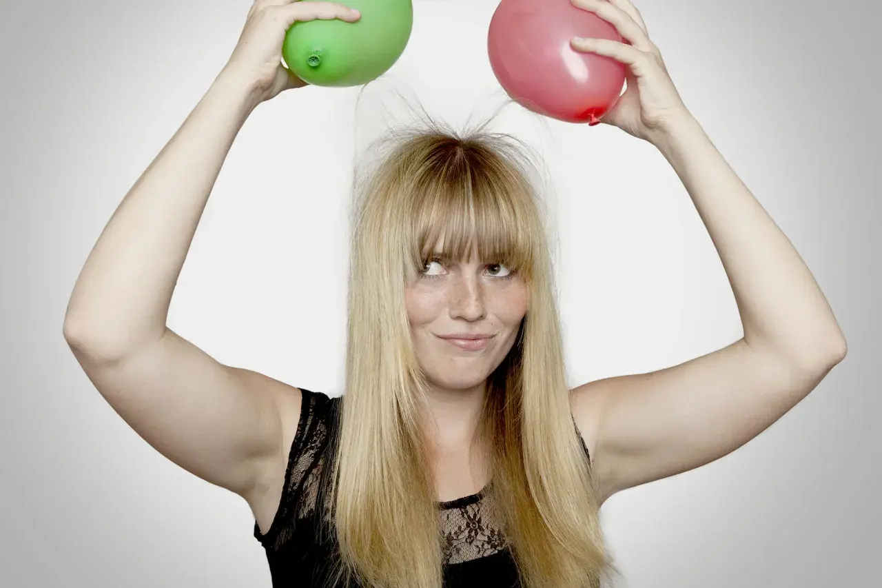 En kvinna med blont hår och fräknar håller upp en grön och en röd ballong ovanför huvudet, vilket får hennes hår att resa sig av statiskt elektricitet.