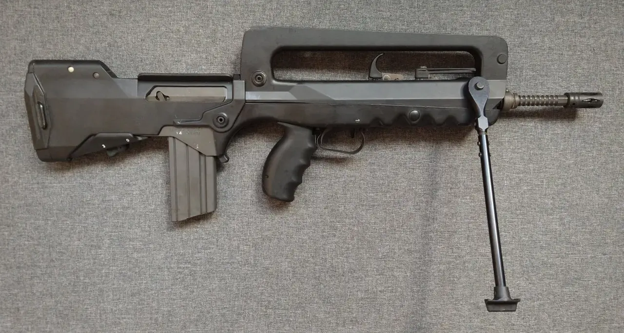 Czarny karabin FAMAS z charakterystycznym uchwytem i dwójnogiem, leżący na szarej tkaninie.