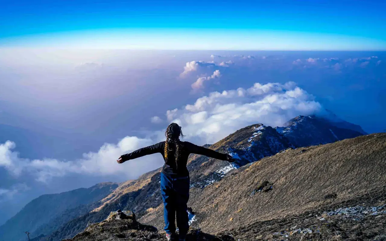 Eine Frau steht auf einem Berggipfel, die Arme ausgebreitet, und blickt auf eine Wolkendecke. Die Frage "wie hoch ist der Kilimandscharo?" hallt in der Weite wider.