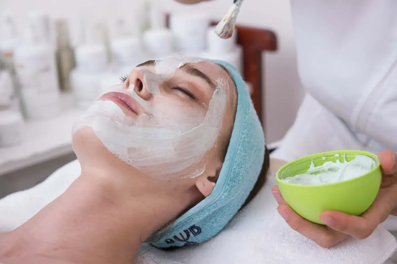 Une femme reçoit un masque ou gommage en premier, appliqué au pinceau sur son visage. Elle est détendue, les yeux fermés.