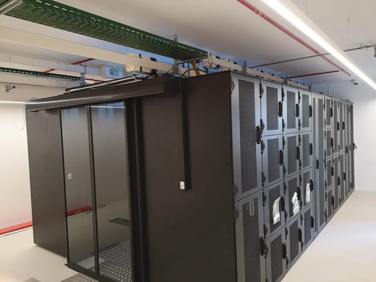 Un micro data center avec des baies de serveurs noires et une porte vitrée. Des câbles et des conduits sont visibles au plafond.