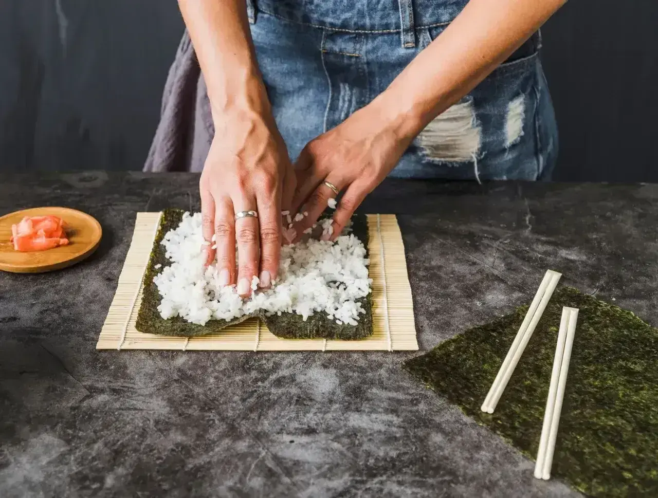 Ręce rozprowadzają ryż na nori, przygotowując zaprawę do sushi. Obok leżą pałeczki i marynowany imbir.