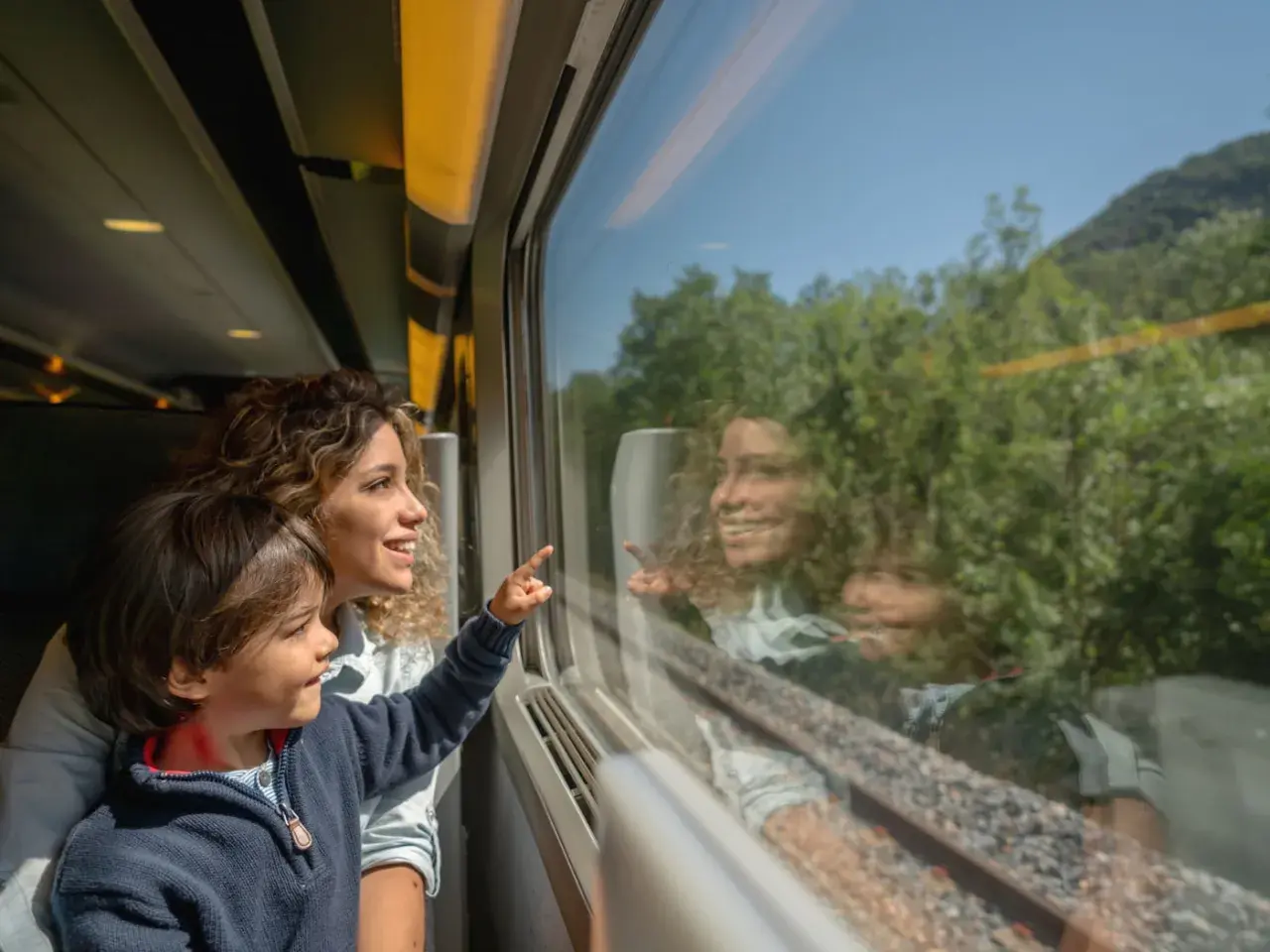 Bambino e madre guardano fuori dal finestrino del treno, indicando qualcosa. Un'ottima idea per i giochi da fare in treno, osservando il paesaggio che scorre.